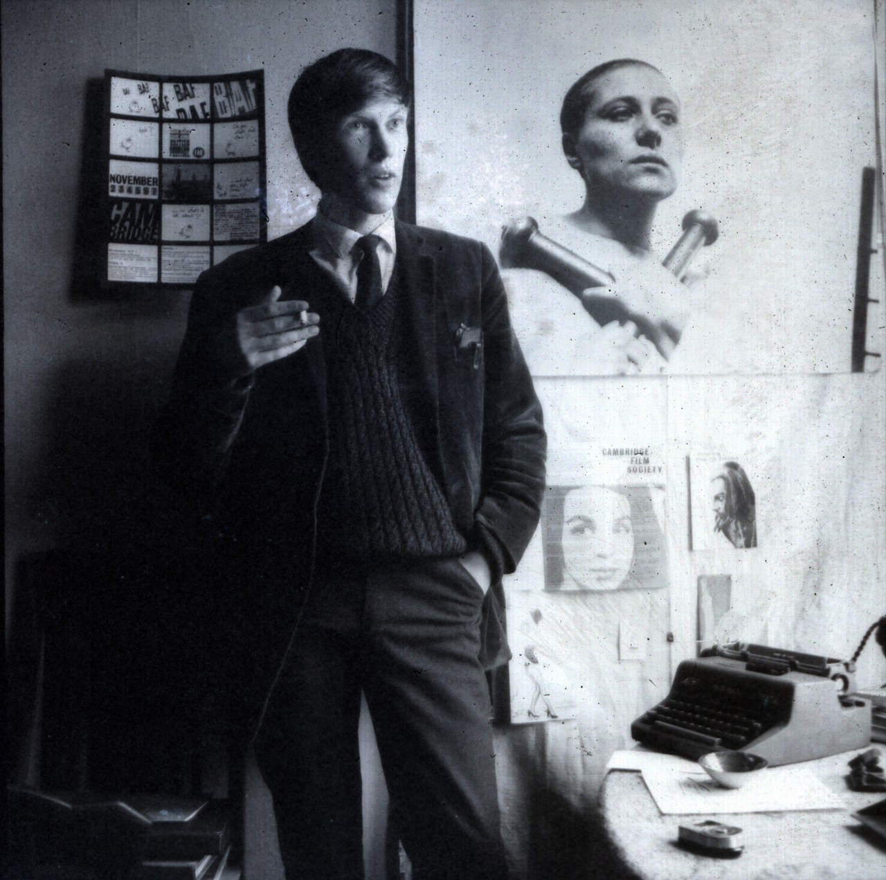 Dick Arnall standing beside a typewriter, with a festival poster and film photographs on the wall behind him