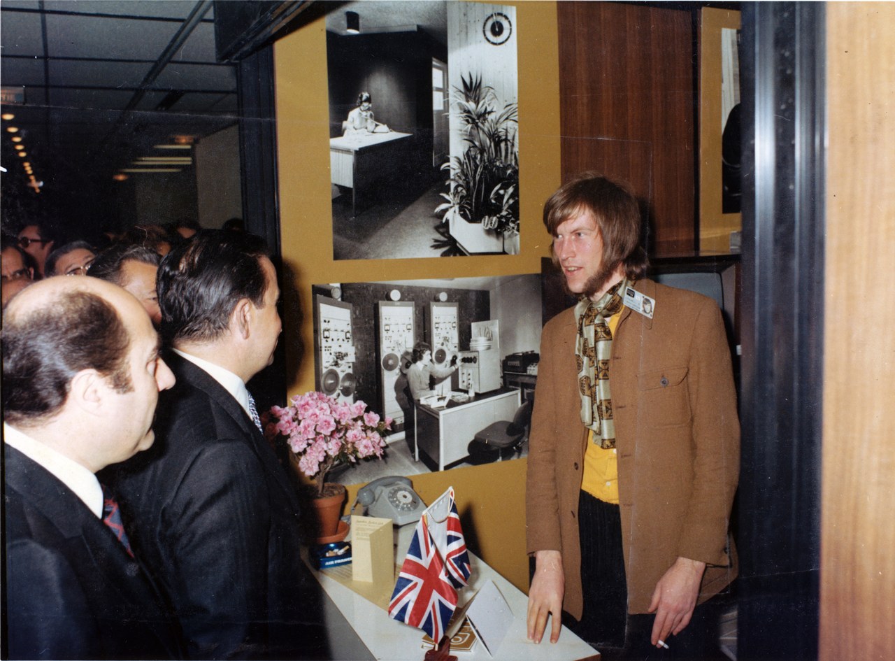 Dick Arnall in a brown jacket and patterned scarf speaking to visitors at a stand displaying film photographs and a small Union Jack